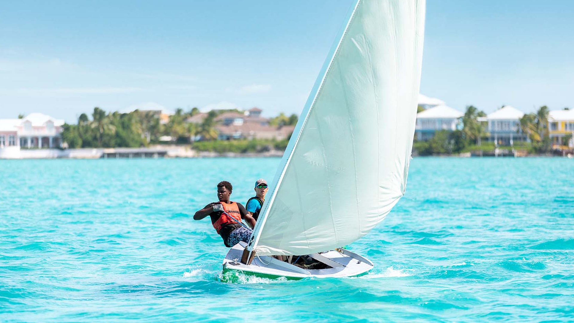 Two young people sailing a small dinghy in turquoise water with palm-lined shore