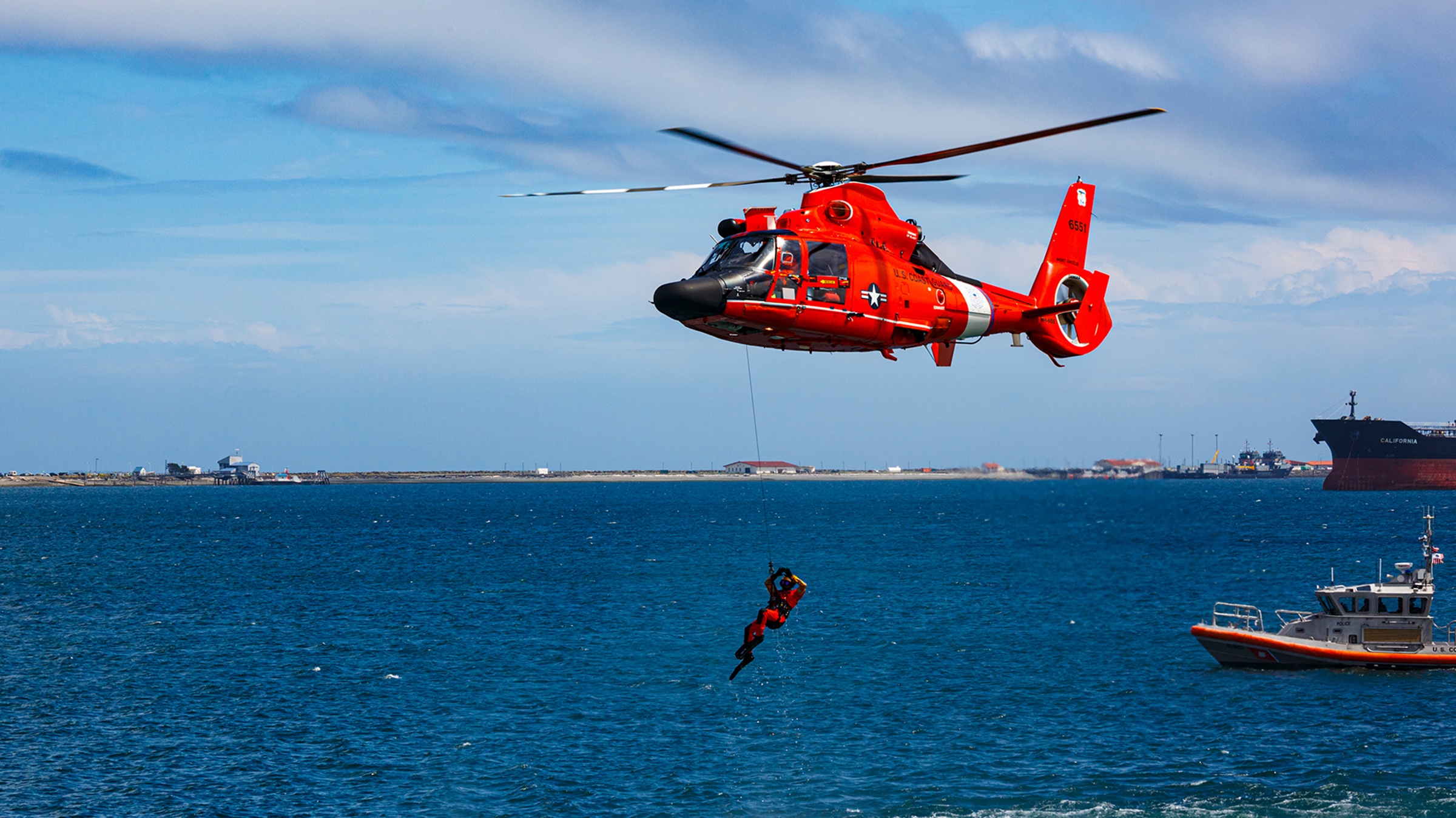 A United States Coast Guard helicopter performing a rescue, with a swimmer suspended on a hoist cable