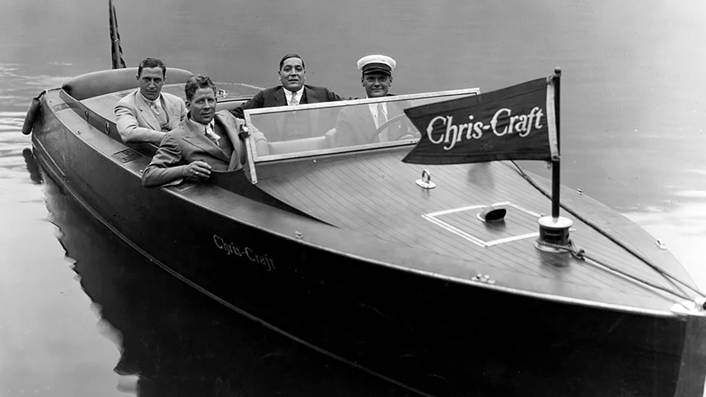 Cuatro hombres de saco y corbata, uno con gorra de capitán, sentados a bordo de una Chris-Craft clásica, foto de época