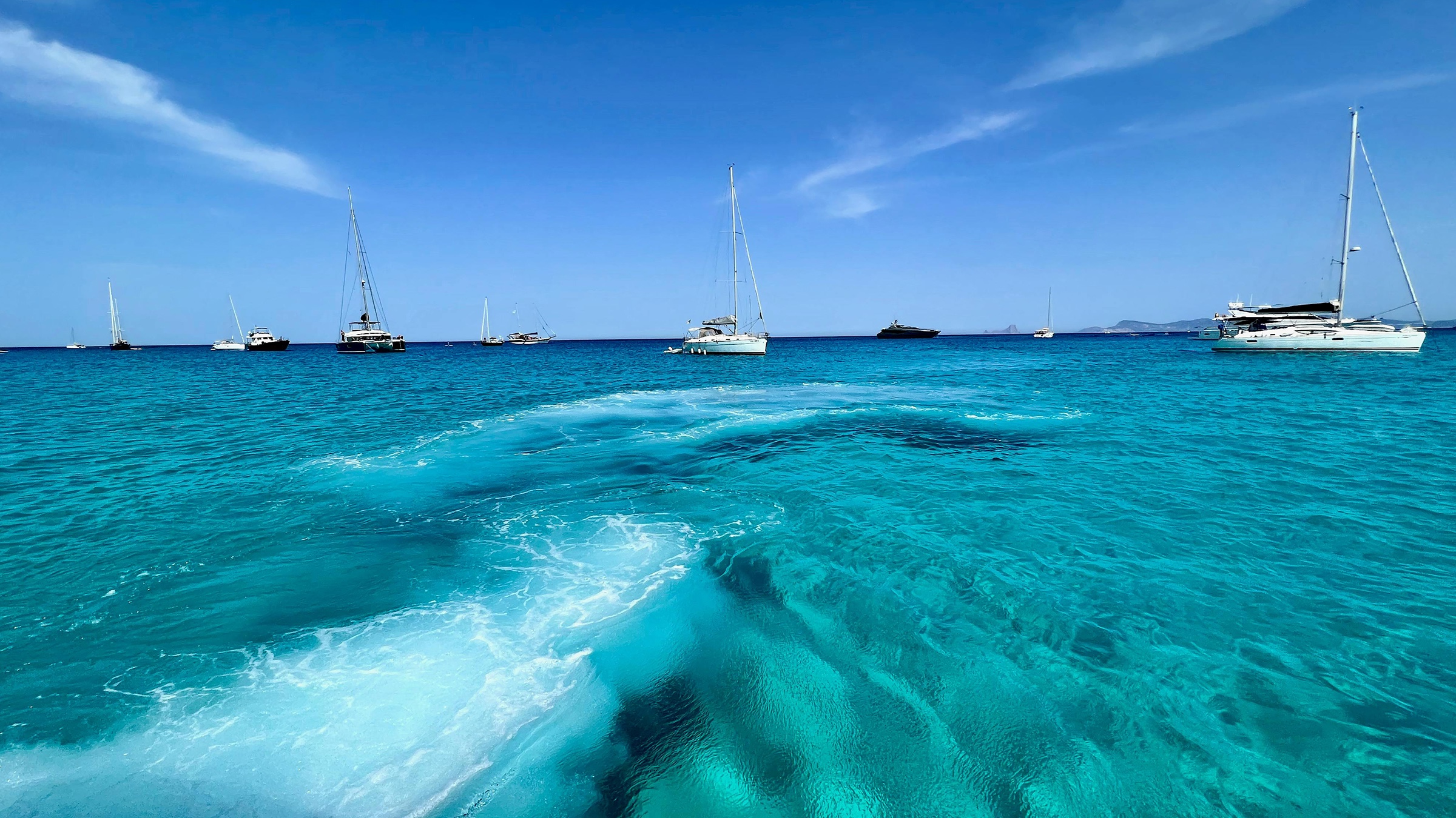 Wake seen from the stern as a yacht leaves a blue anchorage with several boats