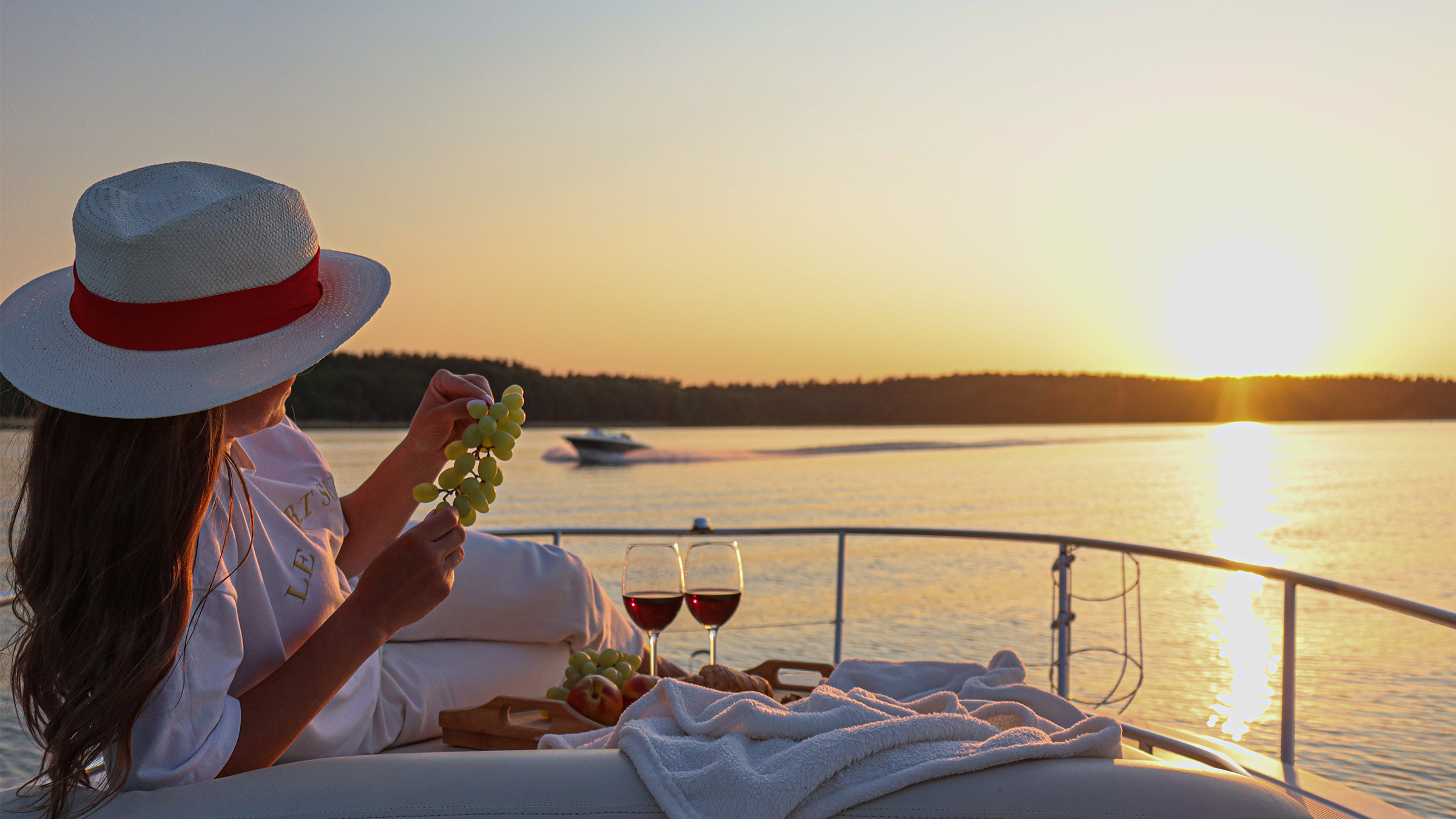 Mujer elegante con sombrero recostada en el lounge de proa con uvas y dos copas de vino al atardecer