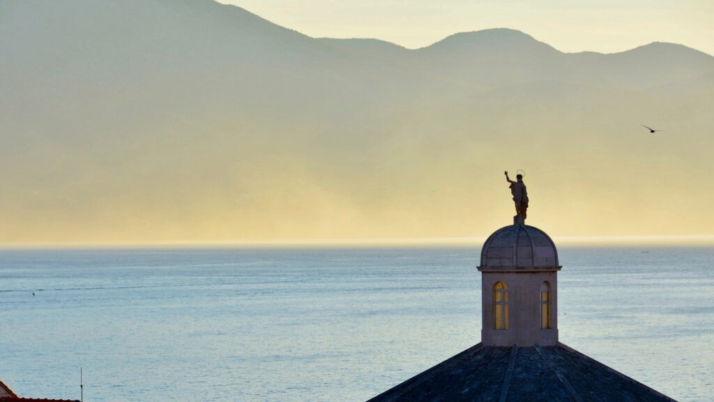 1920 x 1080 Korčula, Croatia Imagen de una cupula con una estatua encima y el atrdecer de fondo sobre un oceano con montañas detrás