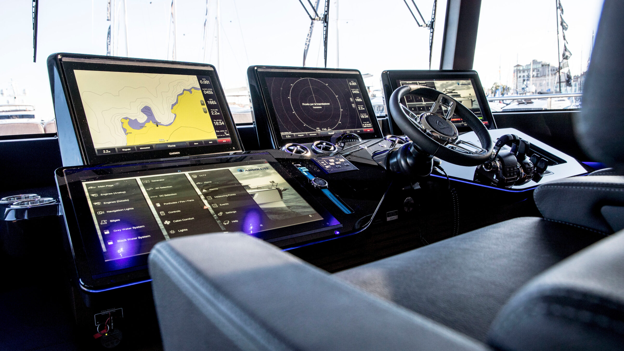 Yacht helm console with several touchscreen navigation displays, a black leather wheel and a marina in the background