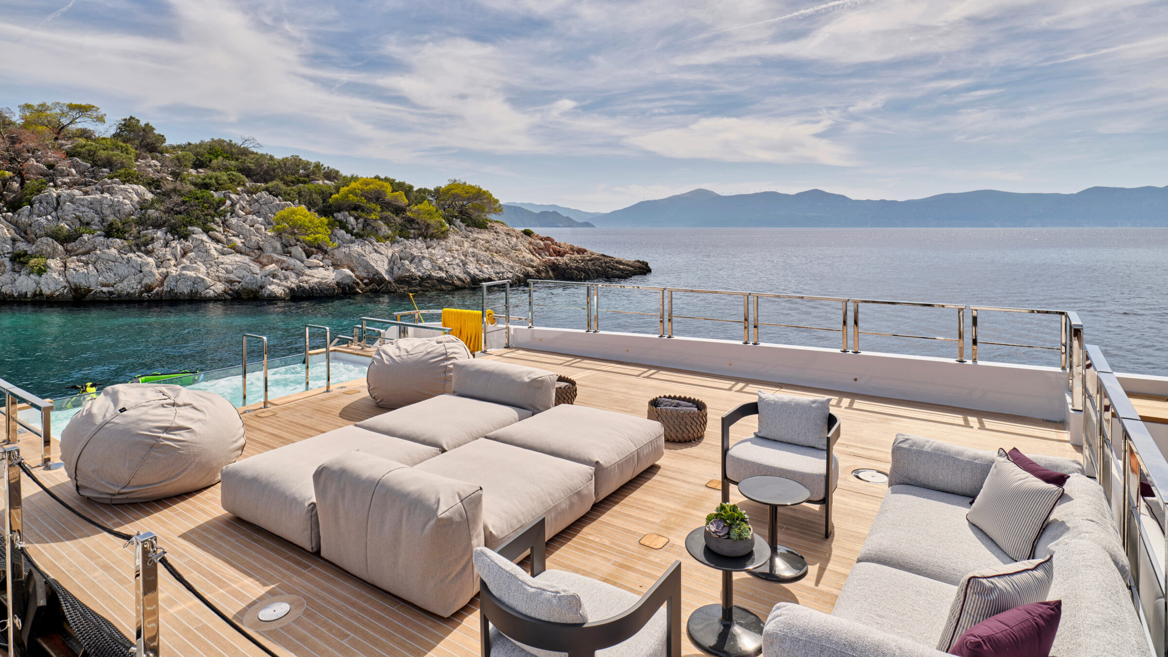 Aft deck of a superyacht with sofas and poufs, at anchor off a rocky coast