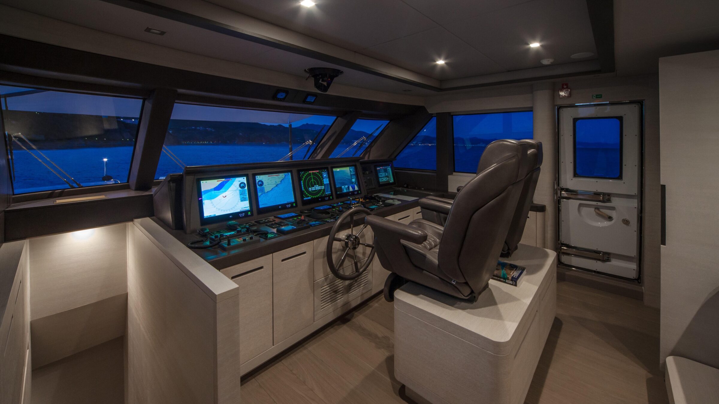 Yacht bridge at night with several navigation displays lit, a steel wheel and leather chairs facing large windows over the coast