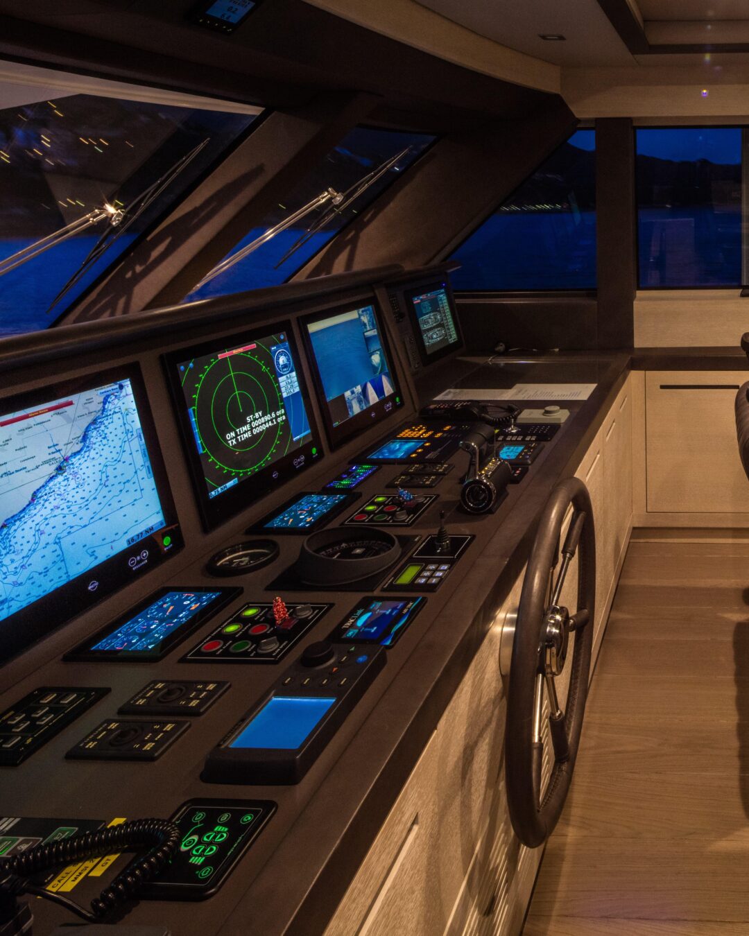 Detail of a bridge console at night with nautical chart and radar displays, illuminated control panels and a steel wheel