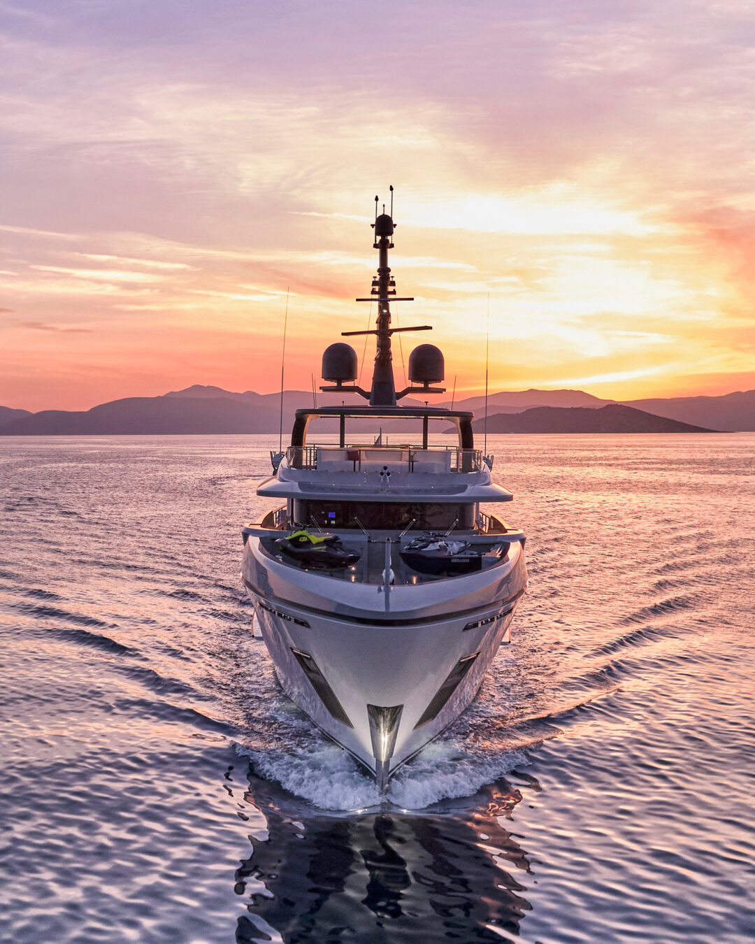 Front view of a superyacht under way at sunset, mountains in the background