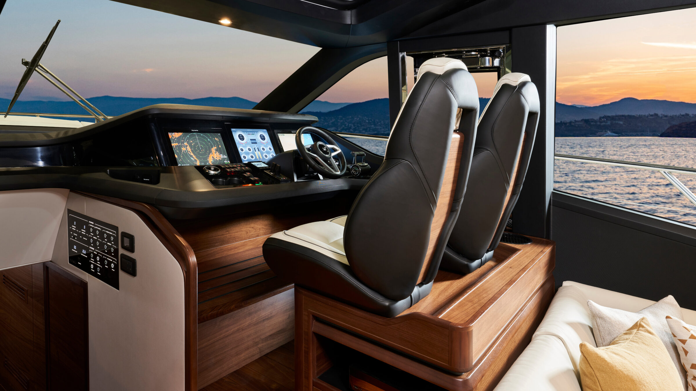 Yacht interior helm with leather seats on a wood base, a console of navigation screens and the coast at sunset