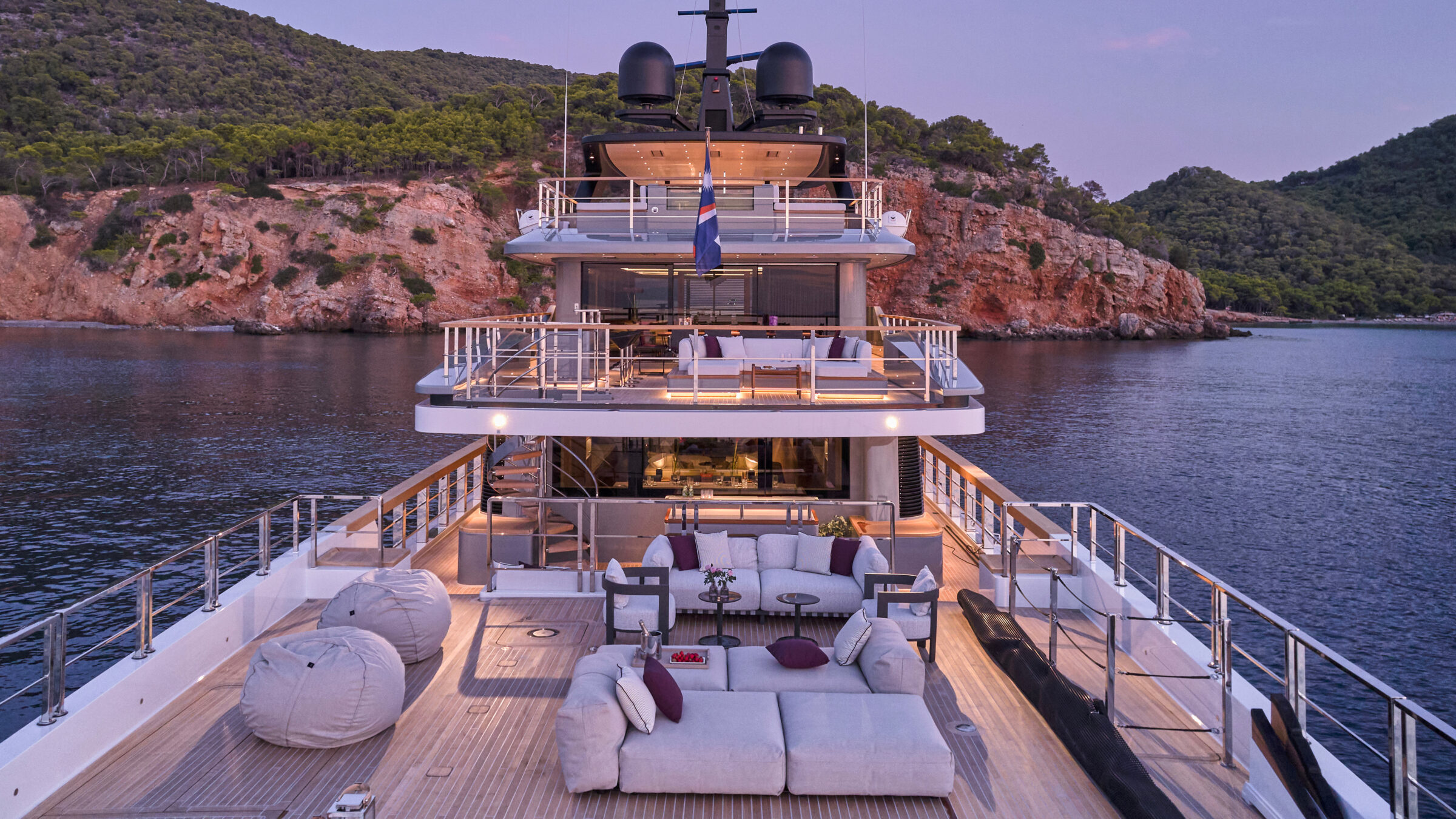 Aft deck of a superyacht at dusk, at anchor in a cove with cliffs